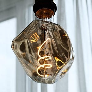 May include: A decorative light bulb with a unique, irregular shape. The bulb has a warm, amber tint and a visible, coiled filament. The bulb is suspended from a black fixture, with a soft, white curtain in the background.