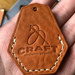 May include: A tan leather tag with a pentagonal shape, featuring a debossed logo of a needle and thread above the text CRAFT and HANDMADE BY YANXU. The tag has a small hole at the top and is stitched along the bottom edges with white thread.