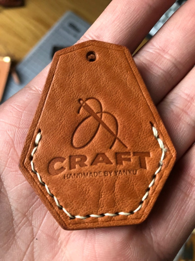 May include: A tan leather tag with a pentagonal shape, featuring a debossed logo of a needle and thread above the text CRAFT and HANDMADE BY YANXU. The tag has a small hole at the top and is stitched along the bottom edges with white thread.