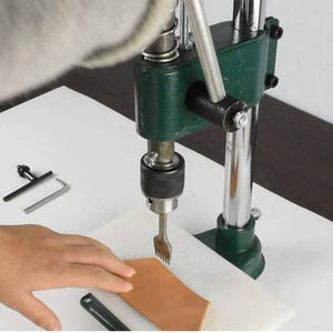 Puede incluir: Una herramienta de punzonado de cuero verde y plateado con una pieza de cuero marrón debajo. La herramienta tiene un mango y un pedal. La herramienta se utiliza para perforar agujeros en el cuero.