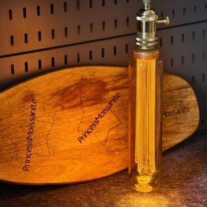 May include: A decorative light bulb with a brass base and a clear glass tube containing vertical filaments. The bulb is next to a wooden oval-shaped board with the text "PrincessMoissanite" and star designs.