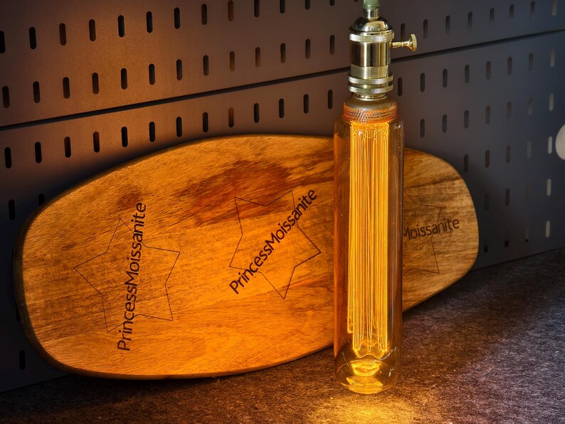 May include: A decorative light bulb with a brass base and a clear glass tube containing vertical filaments. The bulb is next to a wooden oval-shaped board with the text "PrincessMoissanite" and star designs.