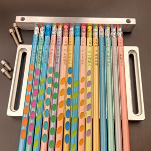 May include: A selection of brightly coloured pencils, each featuring Chinese characters and a polka dot design. The pencils, in various colours, are arranged in a row and rest on a silver metal stand. The pencils are approximately 19 cm long.