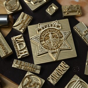 May include: A collection of brass metal stamps for leather crafting, including a large star-shaped badge stamp with the text DEPUTY SHERIFF'S OFFICE SAN MATEO CO, alongside various rectangular stamps with engraved letters and symbols on a dark leather piece.