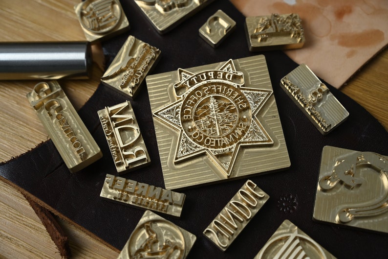 May include: A collection of brass metal stamps for leather crafting, including a large star-shaped badge stamp with the text DEPUTY SHERIFF'S OFFICE SAN MATEO CO, alongside various rectangular stamps with engraved letters and symbols on a dark leather piece.