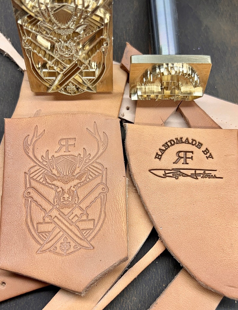 May include: Two brass metal stamps with handles, one featuring a deer head and crossed knives, the other with the text "HANDMADE BY RF" and a signature. Two pieces of tan leather show the stamped impressions of these designs in dark brown ink.