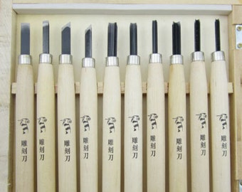 May include: A set of ten wood carving tools with wooden handles. The tools are arranged in a wooden box. The tools have different shaped blades and are labeled in Japanese.