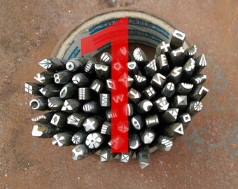 Pode incluir: Uma coleção de carimbos de metal com vários desenhos, incluindo formas geométricas, letras e símbolos. Os carimbos estão dispostos em forma de coração e são mantidos juntos por um rolo de fita.
