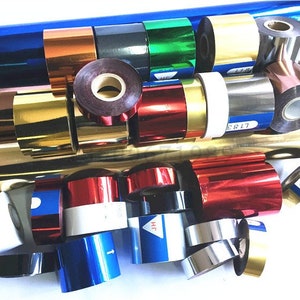 May include: Assortment of colorful rolls of foil ribbon in various colors, including gold, silver, red, blue, green, and black. The rolls are arranged in a pile on a white surface.