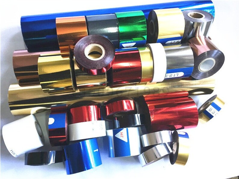 May include: Assortment of colorful rolls of foil ribbon in various colors, including gold, silver, red, blue, green, and black. The rolls are arranged in a pile on a white surface.