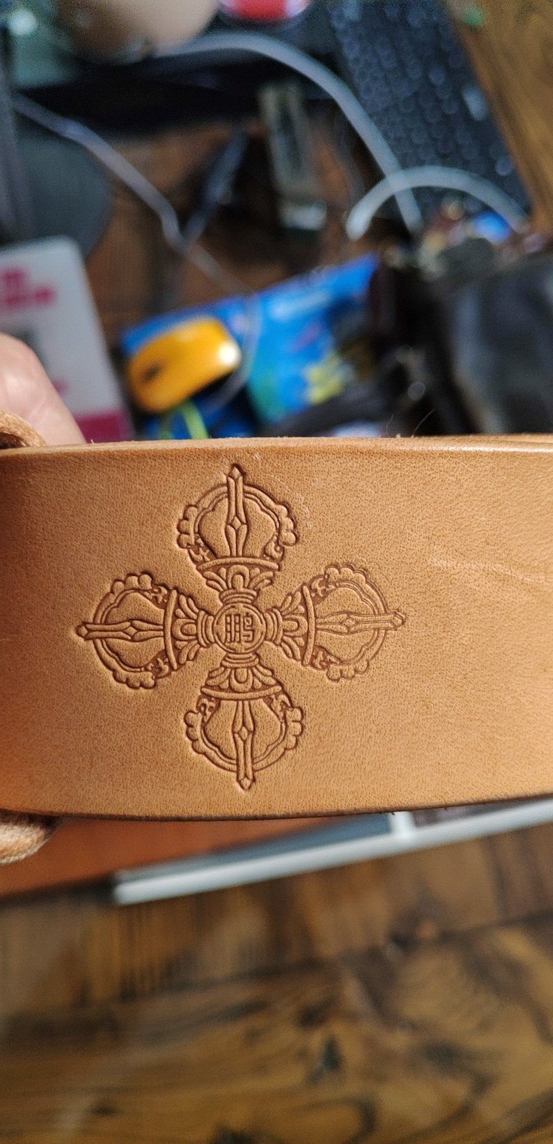 May include: A close-up of a light brown leather belt with a decorative embossed design of a stylized symbol with four arms and a central circle. The symbol is in a light brown color.