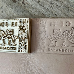 May include: A rectangular brass metal stamp featuring a relief design of two tigers, a column of intricate characters, and the text HANANECHEII. Beside the stamp is a leather surface showing a clear, debossed impression of the same tiger design.