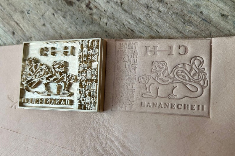 May include: A rectangular brass metal stamp featuring a relief design of two tigers, a column of intricate characters, and the text HANANECHEII. Beside the stamp is a leather surface showing a clear, debossed impression of the same tiger design.