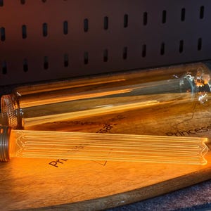 May include: Two Edison-style light bulbs with warm, glowing filaments rest on a wooden cutting board. One bulb is fully assembled with a brass base, while the other is a clear glass tube. The board has text printed on it.