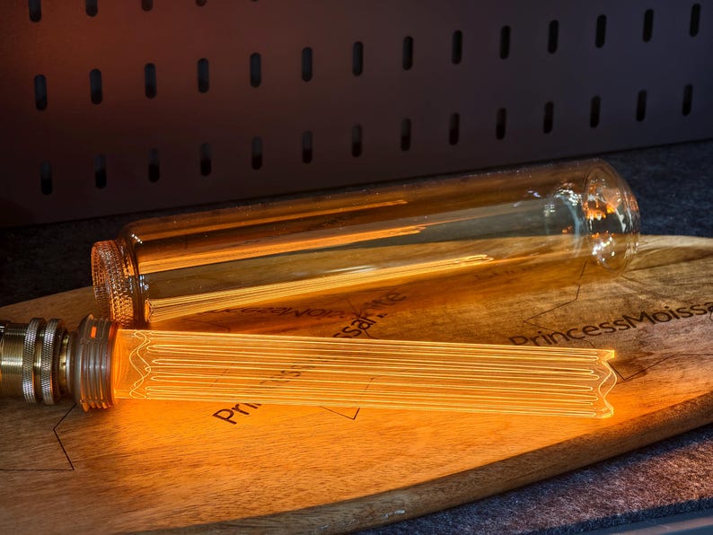 May include: Two Edison-style light bulbs with warm, glowing filaments rest on a wooden cutting board. One bulb is fully assembled with a brass base, while the other is a clear glass tube. The board has text printed on it.