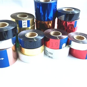 May include: Assortment of colorful ribbon rolls, including black, gold, red, blue, and silver. The rolls are stacked and arranged on a white surface. Some rolls have text printed on them, such as "83" and "19".