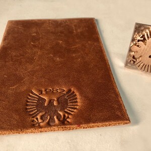 May include: A brown leather piece with a stamped design of a bird with wings spread and the year 1982. The stamp is also shown separately.