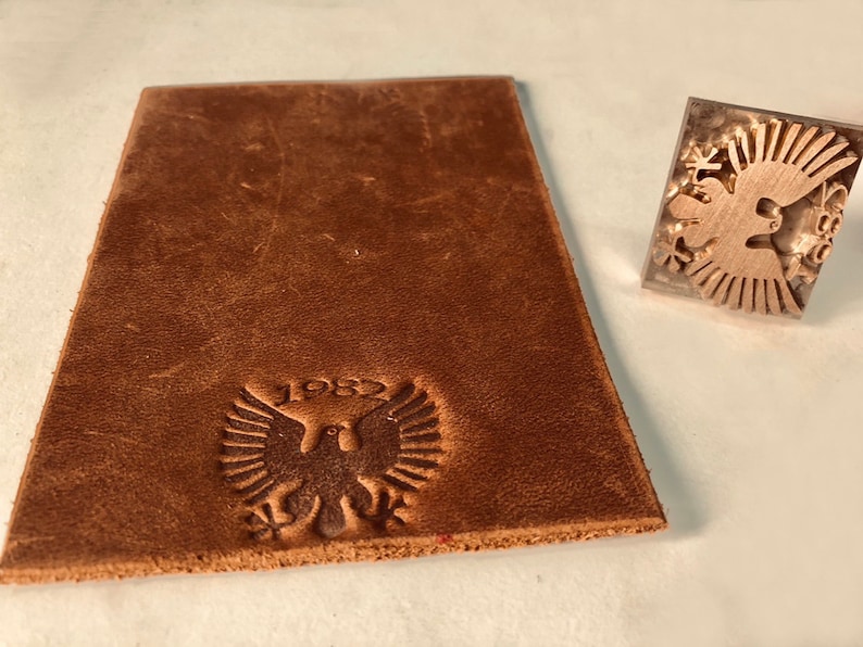 May include: A brown leather piece with a stamped design of a bird with wings spread and the year 1982. The stamp is also shown separately.