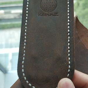May include: A dark brown leather pouch with a rounded top and white stitching along the edges. The surface features a circular embossed logo with an intricate knotwork pattern above the text KevinZ, centered on the front of the leather item.
