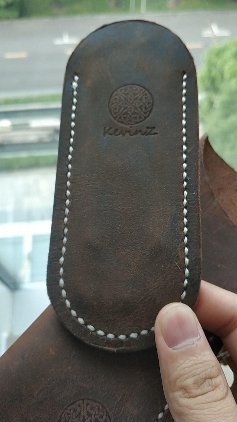 May include: A dark brown leather pouch with a rounded top and white stitching along the edges. The surface features a circular embossed logo with an intricate knotwork pattern above the text KevinZ, centered on the front of the leather item.