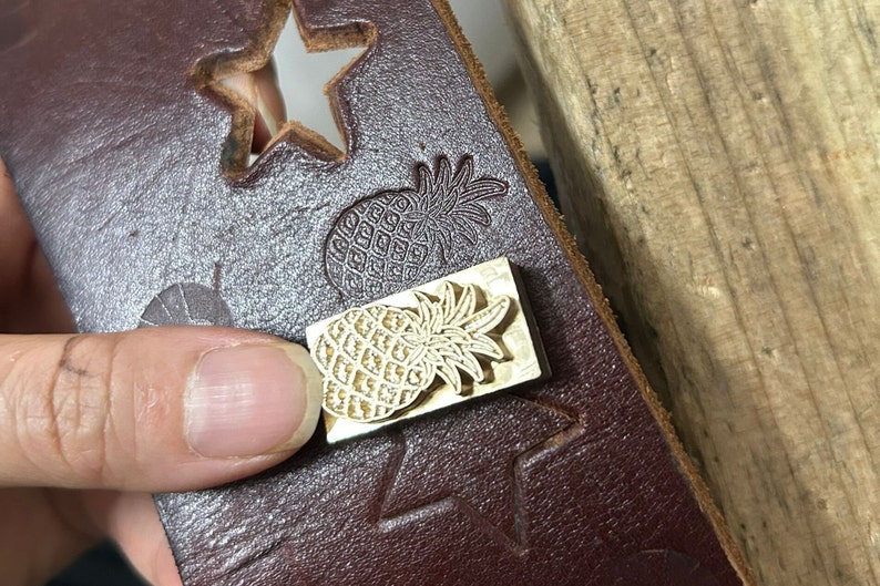 May include: A rectangular brass metal stamp featuring a pineapple design, positioned on a dark brown leather strip. The leather strip displays a cut-out star shape and a stamped pineapple impression, showing the tool's function for leather crafting.