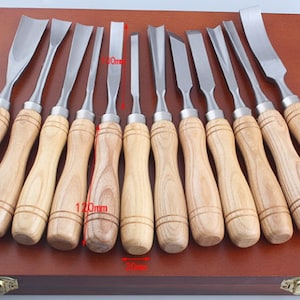 Może przedstawiać: Zestaw 12 dłut do rzeźbienia w drewnie z drewnianymi uchwytami, ułożonych w rzędzie na drewnianej powierzchni. Dłuta są różnych rozmiarów i kształtów, a wszystkie są ostre. Dłuta znajdują się w drewnianym pudełku z mosiężnym zatrzaskiem.