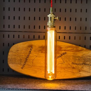 May include: A vintage-style pendant light with a long, rectangular Edison bulb. The bulb hangs from a brass fixture and a red cord. It is mounted above a wooden, star-shaped plaque with the text "PrincessMoissanite".
