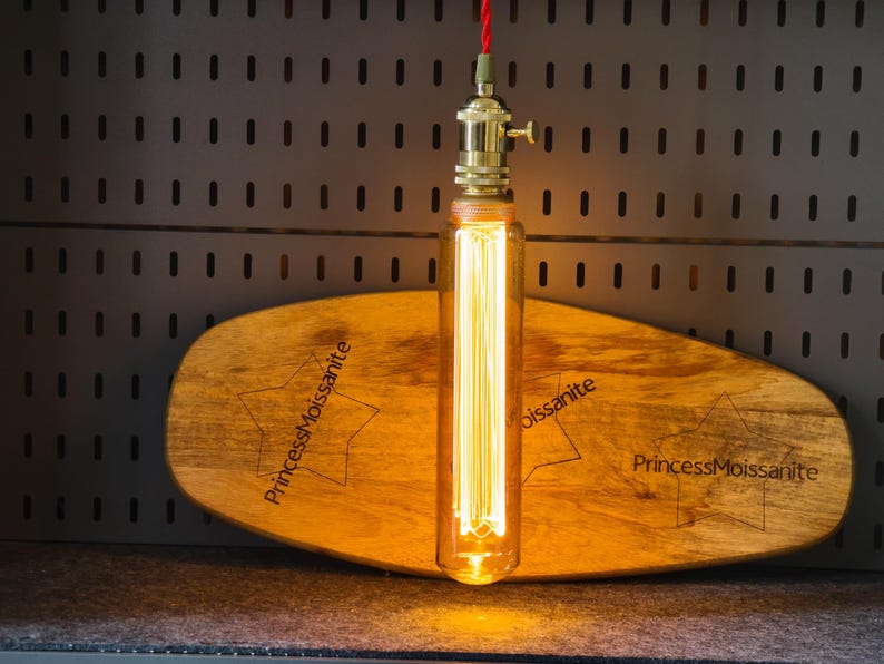 May include: A vintage-style pendant light with a long, rectangular Edison bulb. The bulb hangs from a brass fixture and a red cord. It is mounted above a wooden, star-shaped plaque with the text "PrincessMoissanite".