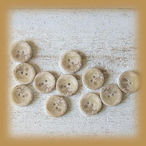 May include: A collection of eleven round, two-hole buttons featuring a marbled, beige and tan swirling pattern. Each button has a smooth, circular surface with two centred holes for attachment, displaying a consistent earthy colour palette.