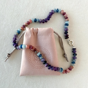 May include: A beaded necklace with a variety of colorful gemstones, including purple, blue, pink, and red. The necklace is displayed next to a pink velvet pouch with the text "Color Spell Necklace". The necklace has a silver clasp.