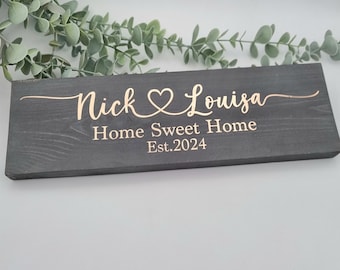 May include: A rectangular, dark gray wooden sign with the names "Nick & Louisa" in a cursive font, joined by a heart symbol. Below, the words "Home Sweet Home Est. 2024" are displayed. The sign is set against a soft, neutral background.