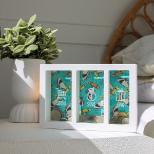May include: White wooden piggy bank with three slots. Each slot has a different design with a shark pattern and a label: Spend, Save, Give.