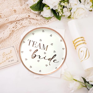 May include: A white plate with a rose gold rim and the words "TEAM Bride" in a script font. The plate is decorated with diamond shapes. A napkin ring and a card with the words "Best wishes to you" are also visible.
