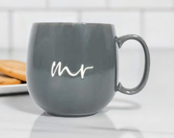 May include: A gray ceramic mug with a white cursive "mr" on the front. The mug has a rounded body and a handle.