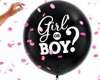 May include: A large, round, black balloon featuring the white text Girl OR BOY? printed on the front. The balloon is surrounded by scattered pink confetti pieces against a plain white background, with a pin held near the surface.