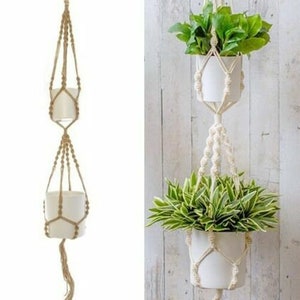 May include: Two white ceramic plant pots hanging from a macrame plant hanger. The macrame is made of natural beige cotton rope. The pots are hanging from a white wall.