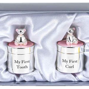 Könnte beinhalten: Zwei silberne Erinnerungsboxen mit rosa Deckeln und Bärenfiguren darauf. Die Boxen sind mit "My First Tooth" und "My First Curl" graviert.
