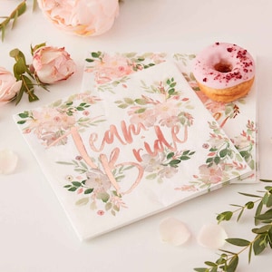 May include: White paper napkins with a floral design and the text "Team Bride" in rose gold foil. The napkins are stacked on a white surface with a pink donut and pink rose petals.