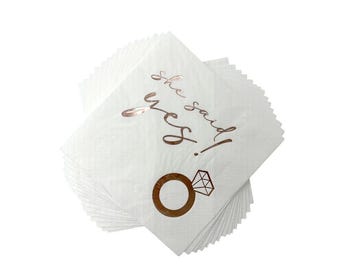 May include: A stack of white paper napkins with a diamond pattern. The napkins have rose gold text that reads "She said yes!" and a rose gold ring graphic. Ideal for a bridal shower or engagement party.