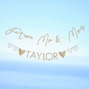 Op de afbeelding: Gouden en witte papieren banner met de tekst "Future Mr & Mrs TAYLOR" en harten. De banner is aan een wit touw geregen.