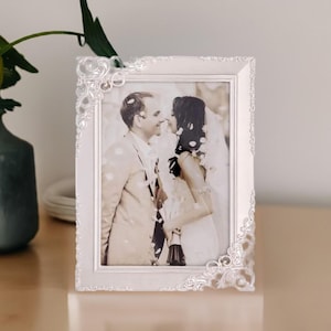 May include: White ornate picture frame with a wedding photo of a couple kissing. The photo is in black and white and shows the couple surrounded by white flower petals.