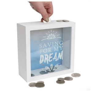 Op de afbeelding: Witte houten spaarpot met een glazen voorzijde. De voorzijde heeft een blauw en wit hemelontwerp met een zon en de tekst "Saving for my dream".