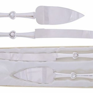 May include: A set of two silver cake knives with heart-shaped handles and clear rhinestones. The knives are in a white box with a cream-colored lining.