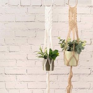 May include: Two macrame plant hangers with a white and natural color. The hangers are hanging from the ceiling and have plants in them. The background is a white brick wall.