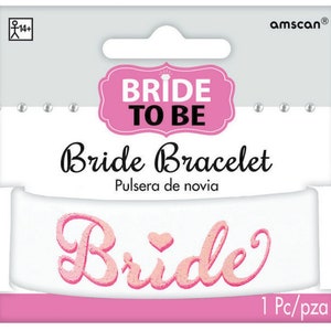 Könnte beinhalten: Weißes "Bride to Be" Armband mit pinkfarbenem Glitzer Schriftzug. Das Armband ist in einer durchsichtigen Plastiktüte verpackt mit einer pinkfarbenen und weißen Kopfkarte, auf der "Bride to Be" und "Bride Bracelet" steht.