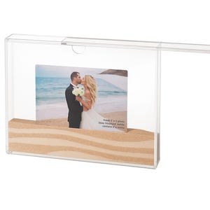 May include: A transparent acrylic photo frame with a beach theme. It holds a 12.7 x 5.08 cm photo and has sand at the base. The photo shows a couple kissing on a beach.
