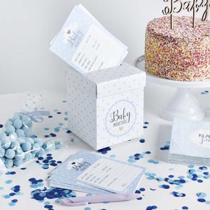 Puede incluir: Una escena de baby shower con un pastel, tarjetas de predicción y una caja de regalo. El pastel está decorado con chispas de colores y un adorno de pastel "Baby". La caja y las tarjetas son de color azul claro con lunares blancos y el texto "Baby Predictions".