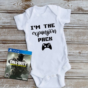 May include: White baby onesie with black text that reads "I'm the expansion pack" and a black video game controller graphic. A video game case for Call of Duty: Infinite Warfare is in the background.