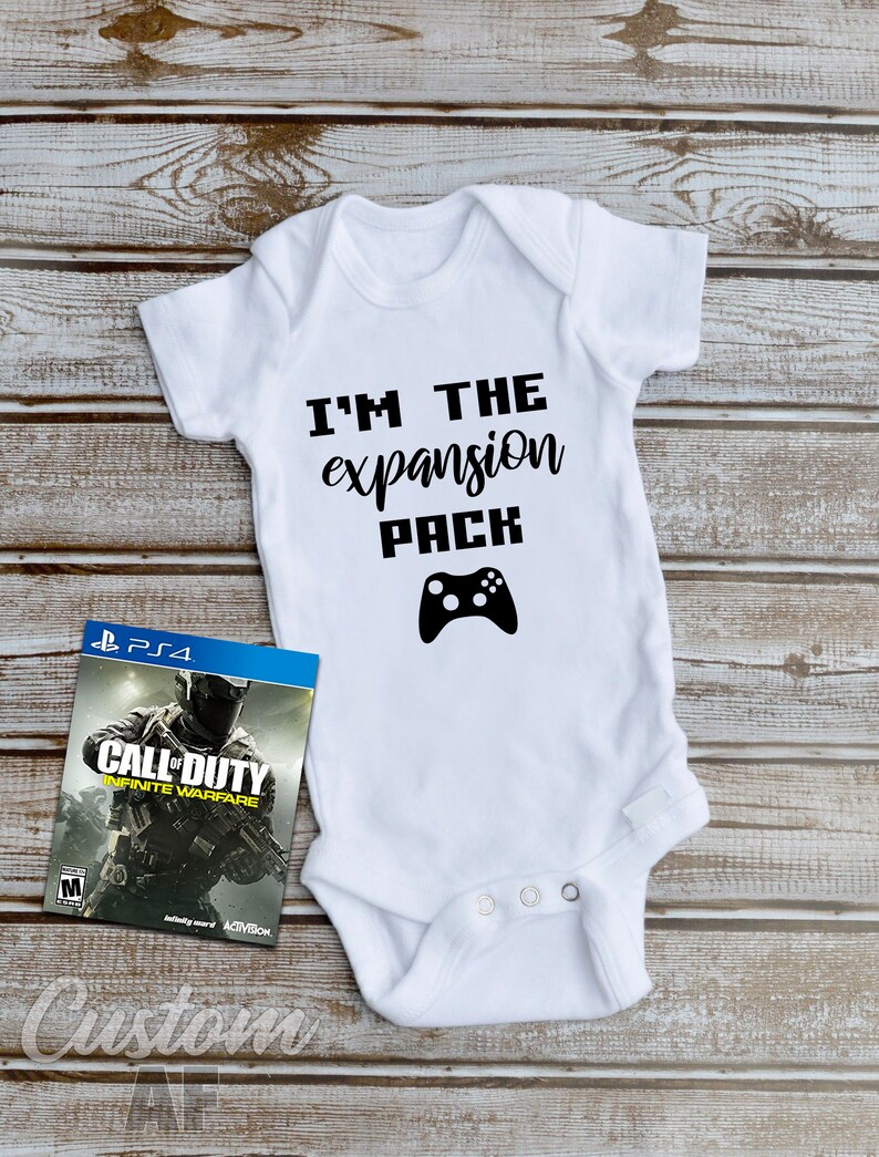 May include: White baby onesie with black text that reads "I'm the expansion pack" and a black video game controller graphic. A video game case for Call of Duty: Infinite Warfare is in the background.