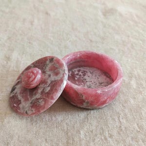 May include: A round pink and white mottled stone trinket box with a matching lid. The lid features a carved rose-shaped handle in the center. The stone has a polished finish with natural veining patterns throughout the circular container and lid.